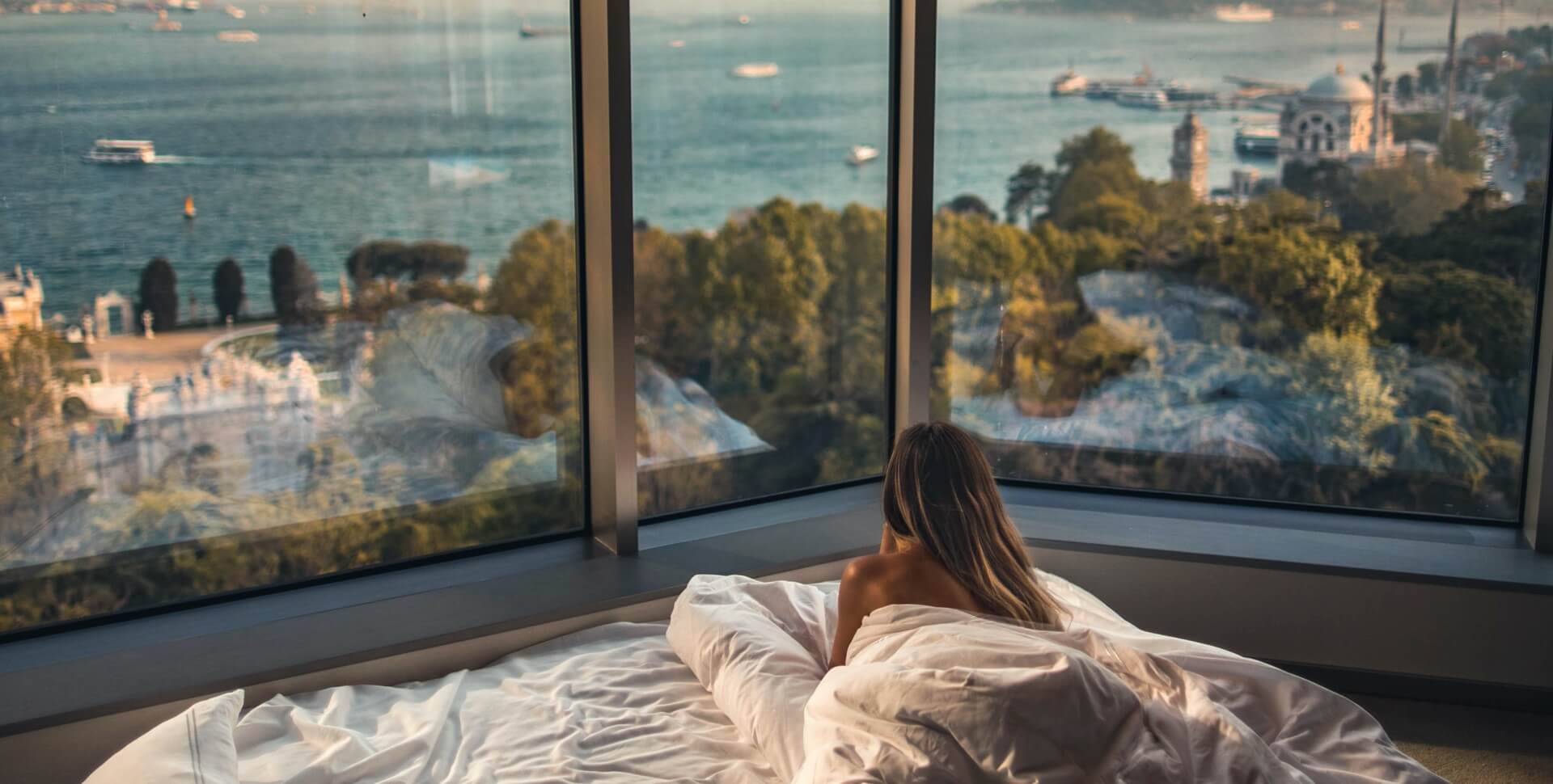 A woman lies on a bed in a room with a large window showing a city view.